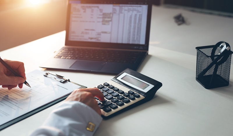 On a white table, a Laptop, Calculator, File pad, and pen holder have been placed, and the businessman is calculating the financial statements of his business.